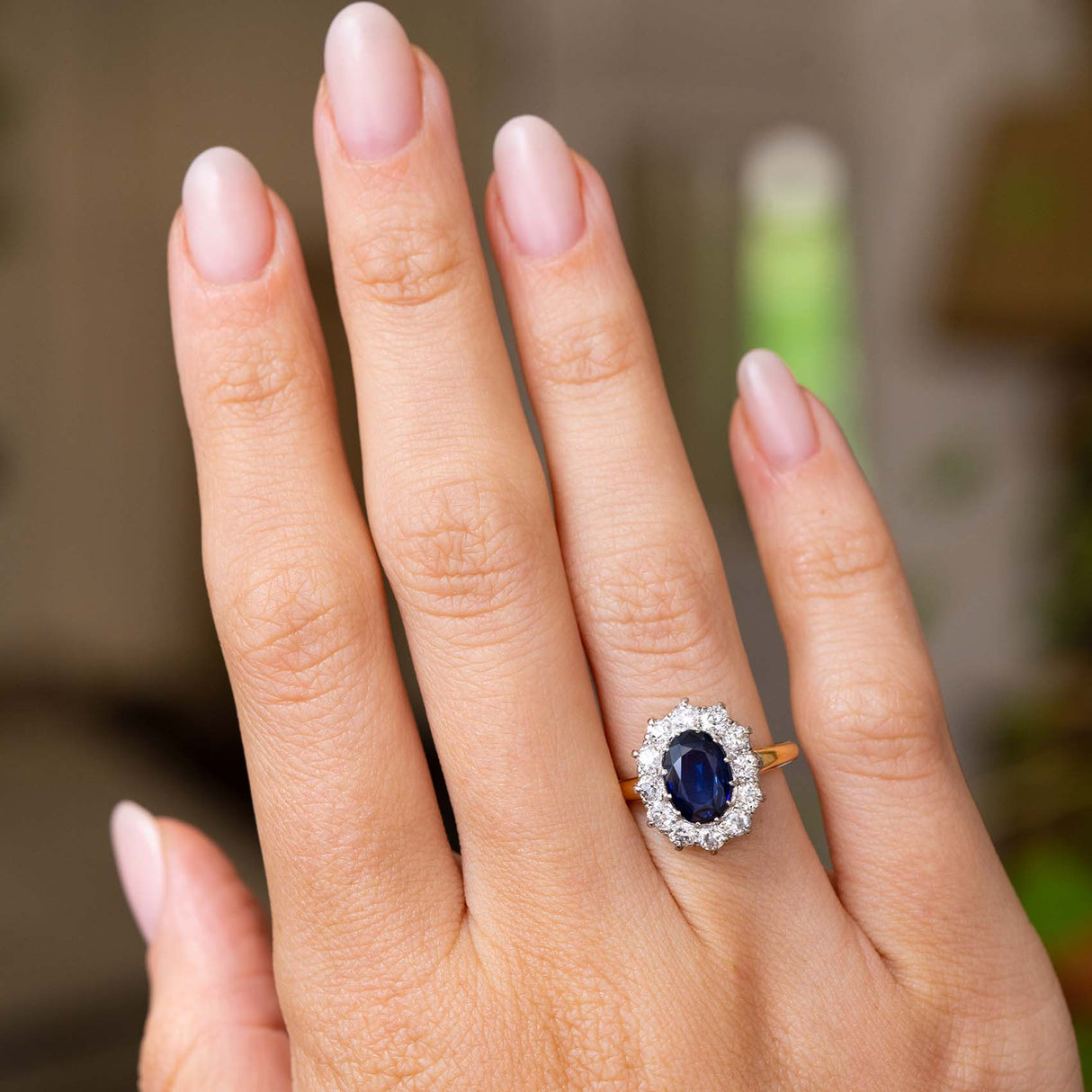 A close-up of a hand wearing a Vintage 1940s cluster engagement ring with a deep oval blue sapphire surrounded by a halo of diamonds, set on a yellow gold band.