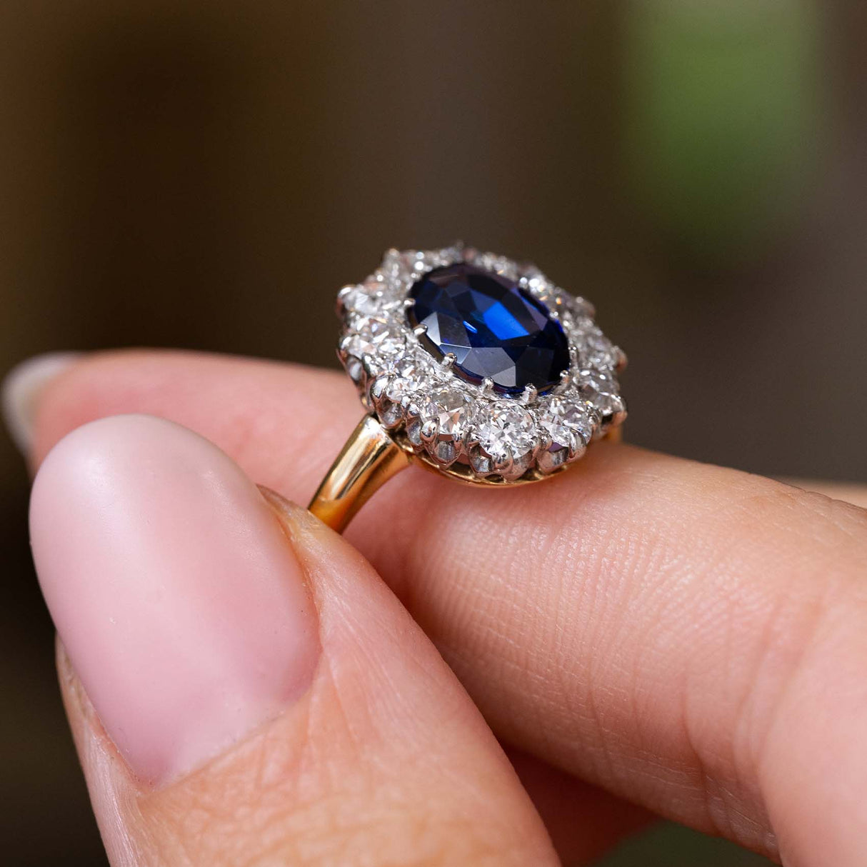 A close up of the side profile and yellow gold band of a Vintage, 1940s blue sapphire and diamond cluster engagement ring worn on a finger.