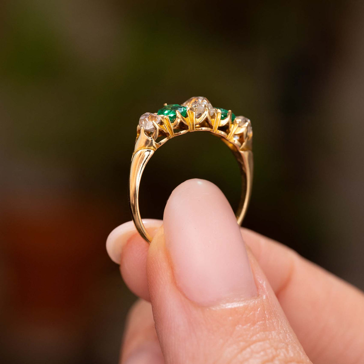 A close-up of a gold ring with three emerald-cut green gemstones and three clear round stones, held between fingers against a blurred dark green background.