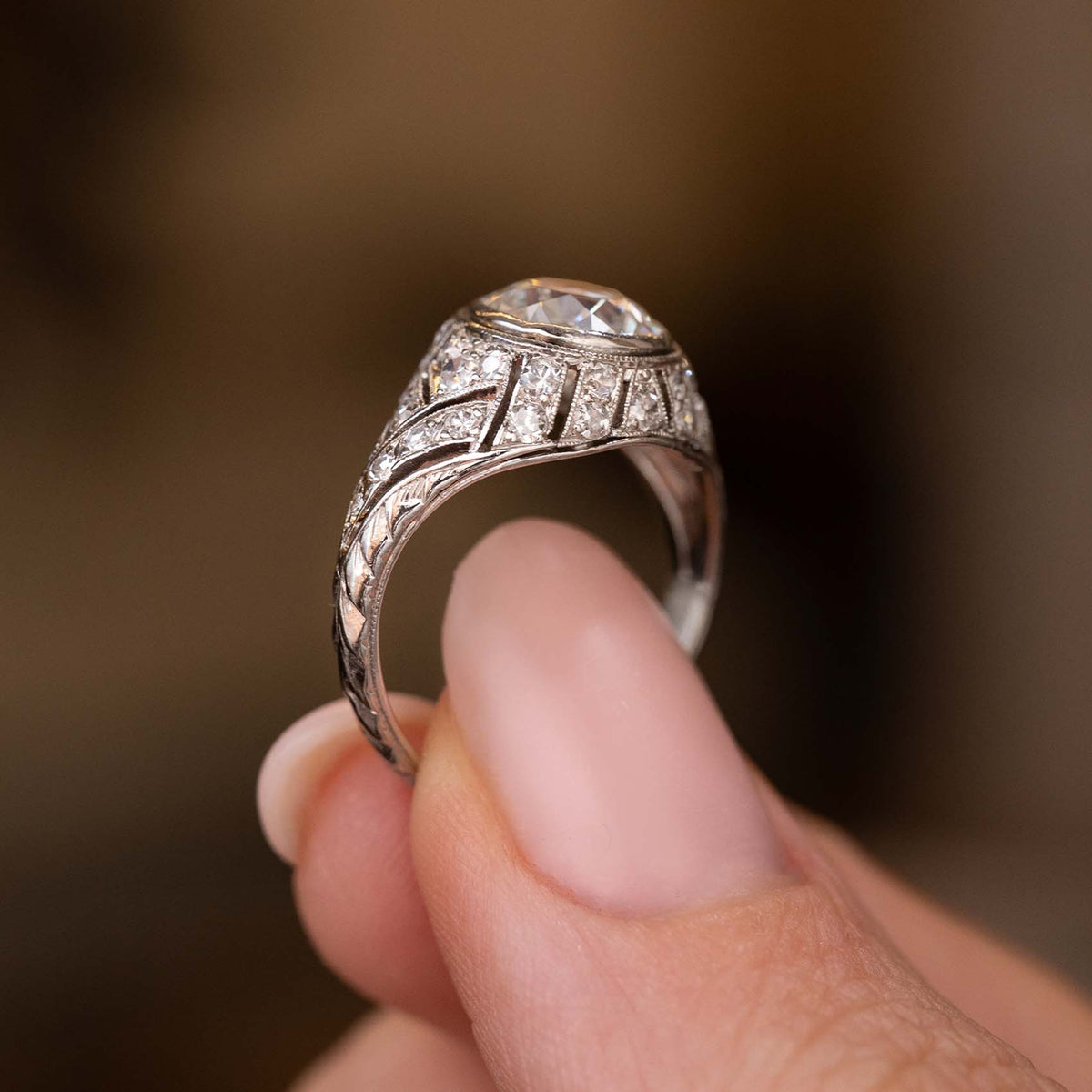 A close-up of a platinum Antique, Art Deco diamond cluster engagement ring held between fingers against a blurred warm brown background.