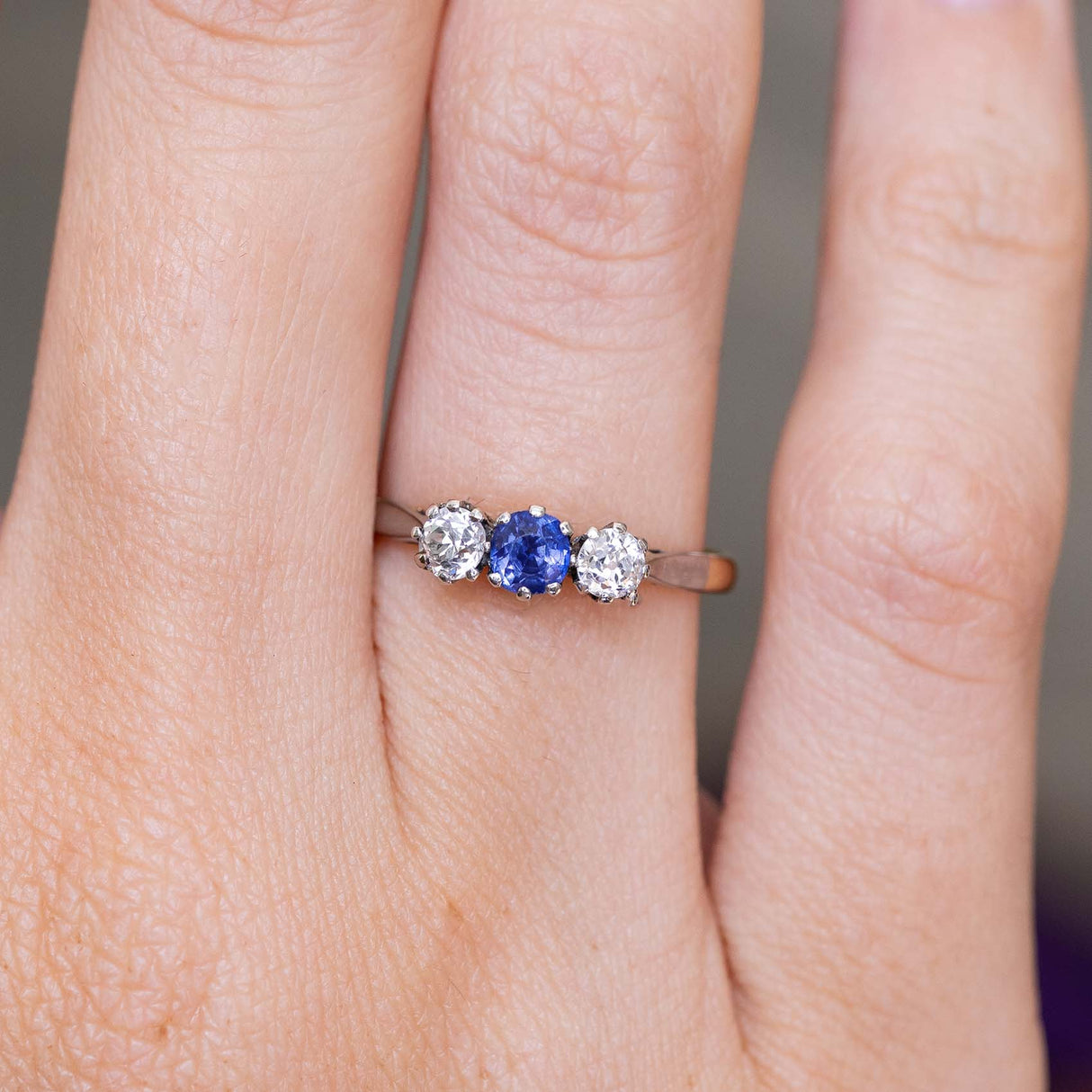 A close-up of an antique trilogy engagement ring worn on a finger, showcasing the central blue sapphire and two diamonds in a platinum setting on a yellow gold band.