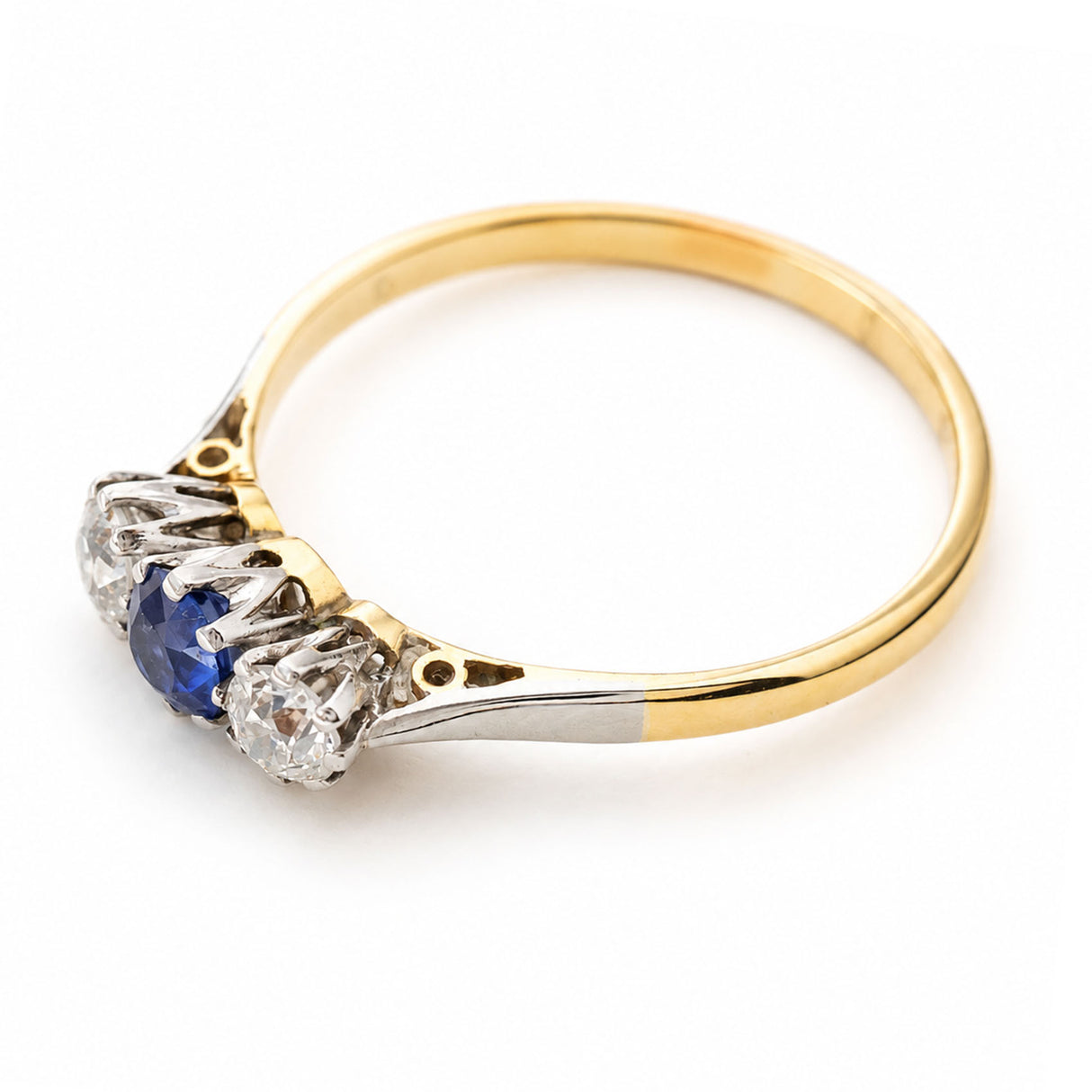 An Antique, Edwardian era blue sapphire and diamond three-stone engagement ring on a yellow gold band shown from above against a white background to showcase the claw-style settings and the details of the shoulders.