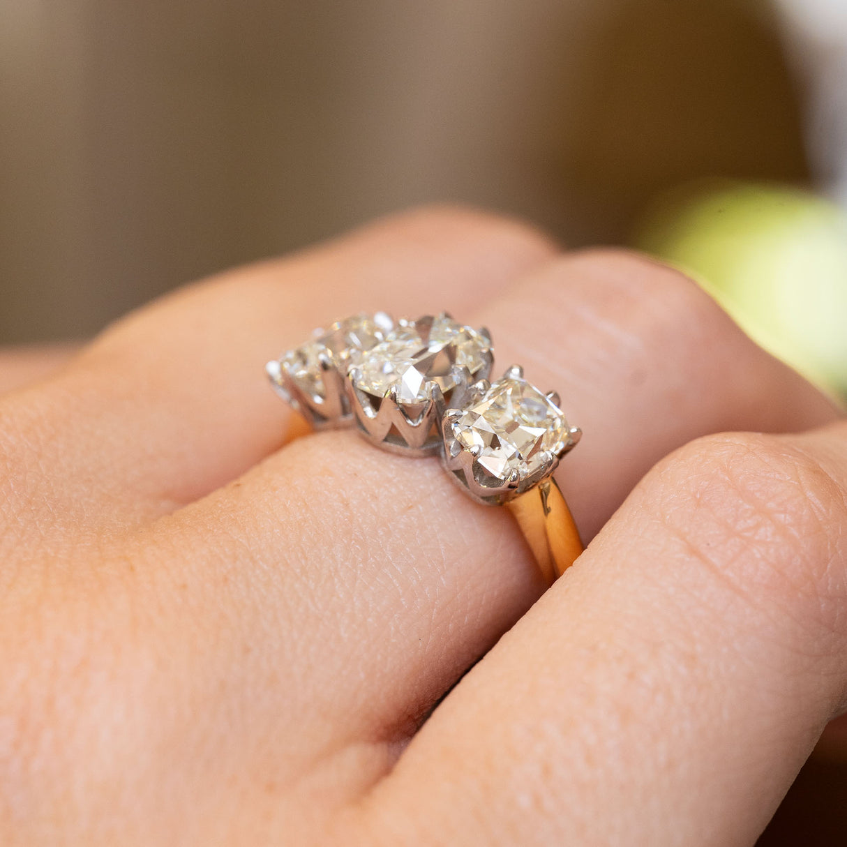 a detailed close up of a vintage diamond engagement ring with three stones on a yellow gold band worn on the hand seen from an angle