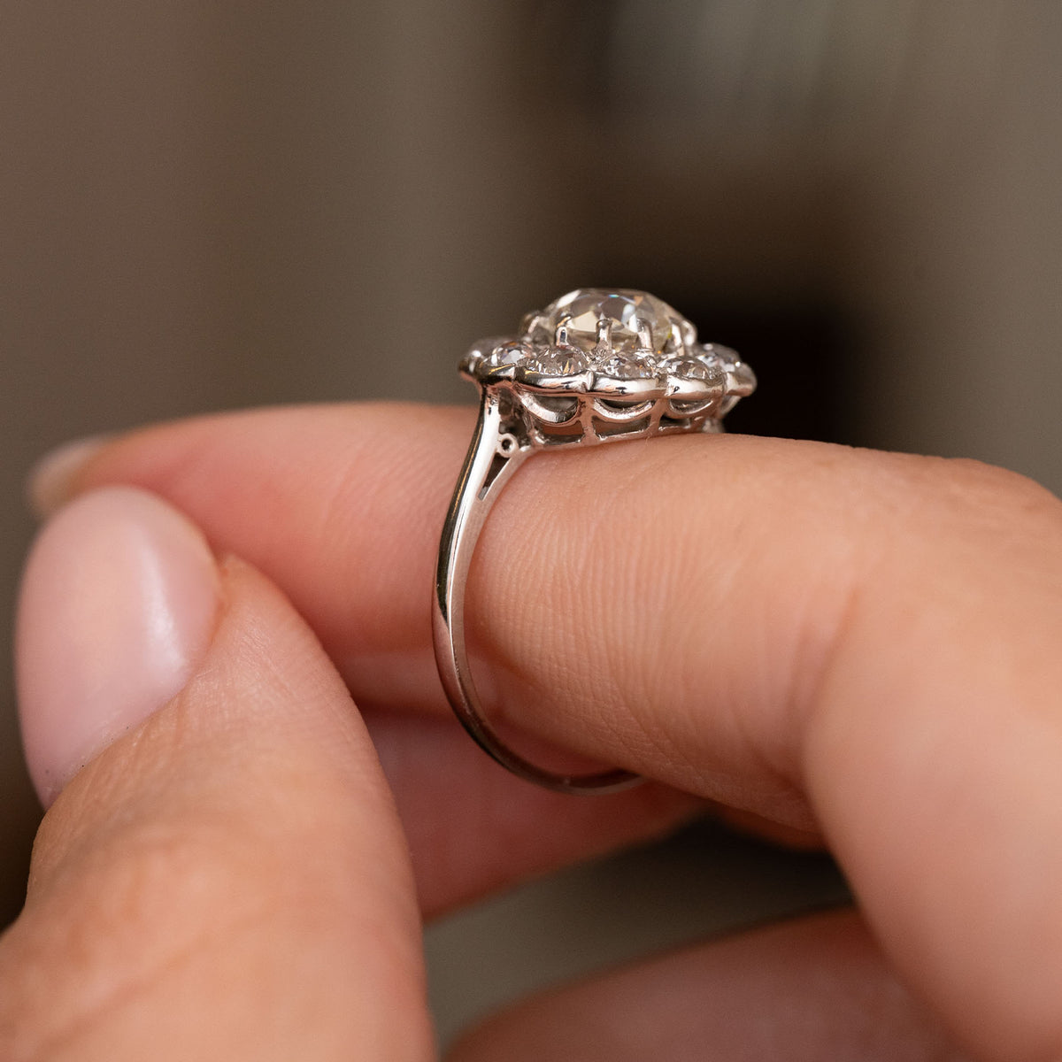 A close-up of a platinum Antique, Edwardian diamond cluster engagement ring shown worn on a person's finger from the side to display the ring's profile.