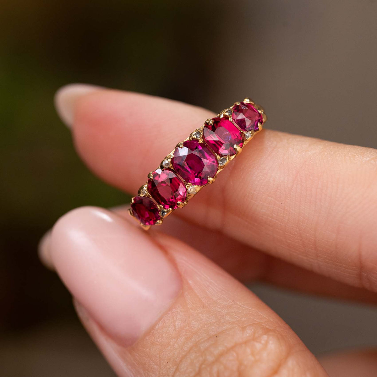 A close-up of a yellow gold Antique, Victorian ruby and diamond five-stone engagement ring held between fingers against a soft, blurred background.