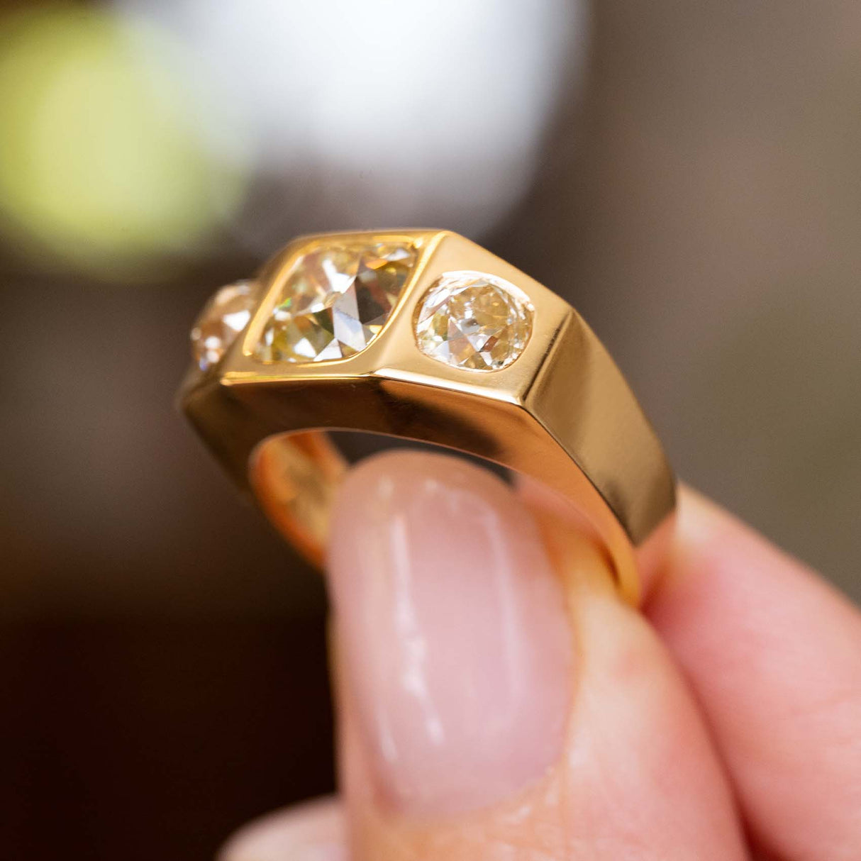 A close-up of a Vintage, 1970s Peruzzi cut diamond three-stone gypsy ring gold ring held between fingers against a blurred dark background.
