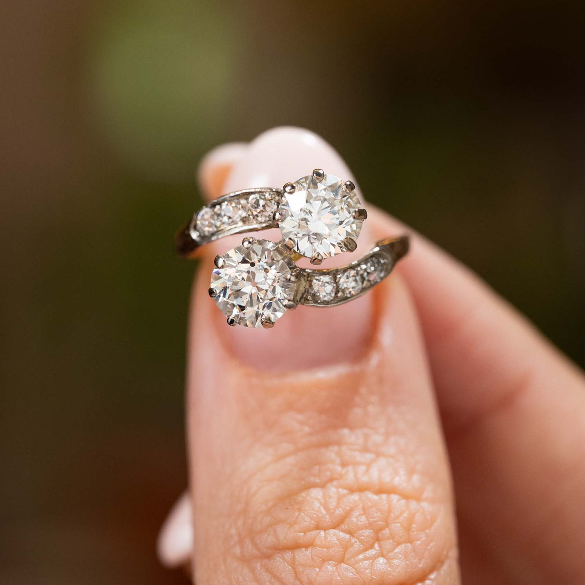 Fingers holding up a diamond two stone engagement ring in yellow gold antique Edwardian held up to the camera to show the details of the diamonds.