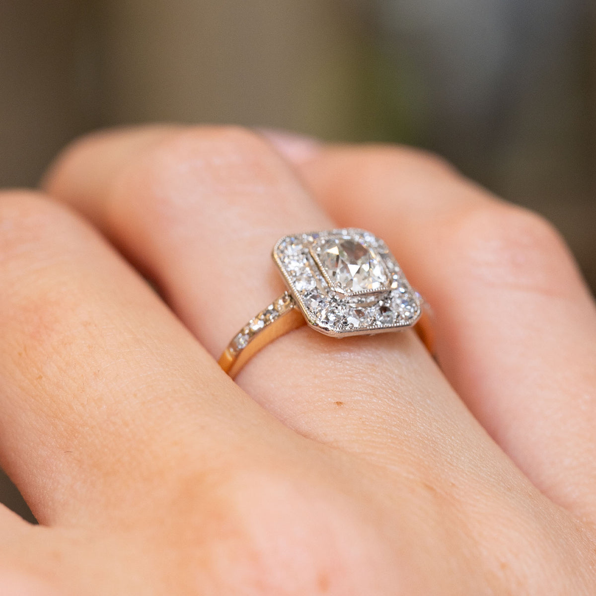 Angled hand view of an Edwardian diamond cluster ring, with a cushion-shaped platinum surround and diamond-set yellow gold shoulders.