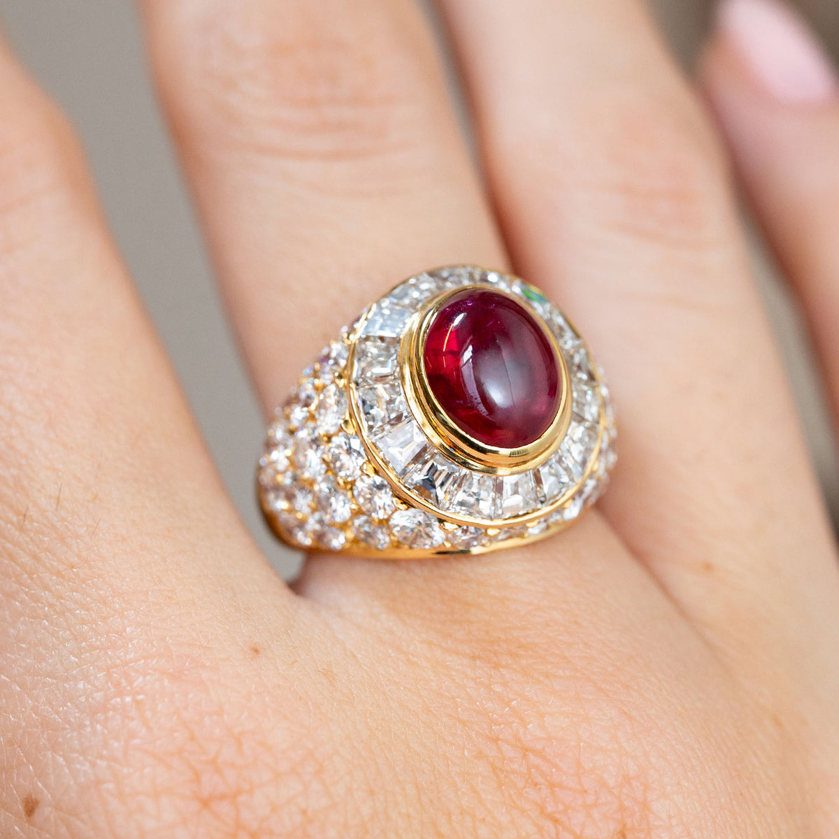 Close-up of the yellow gold bombé ring, featuring an oval ruby framed by tapered step-cut diamonds and pavé-set shoulders.