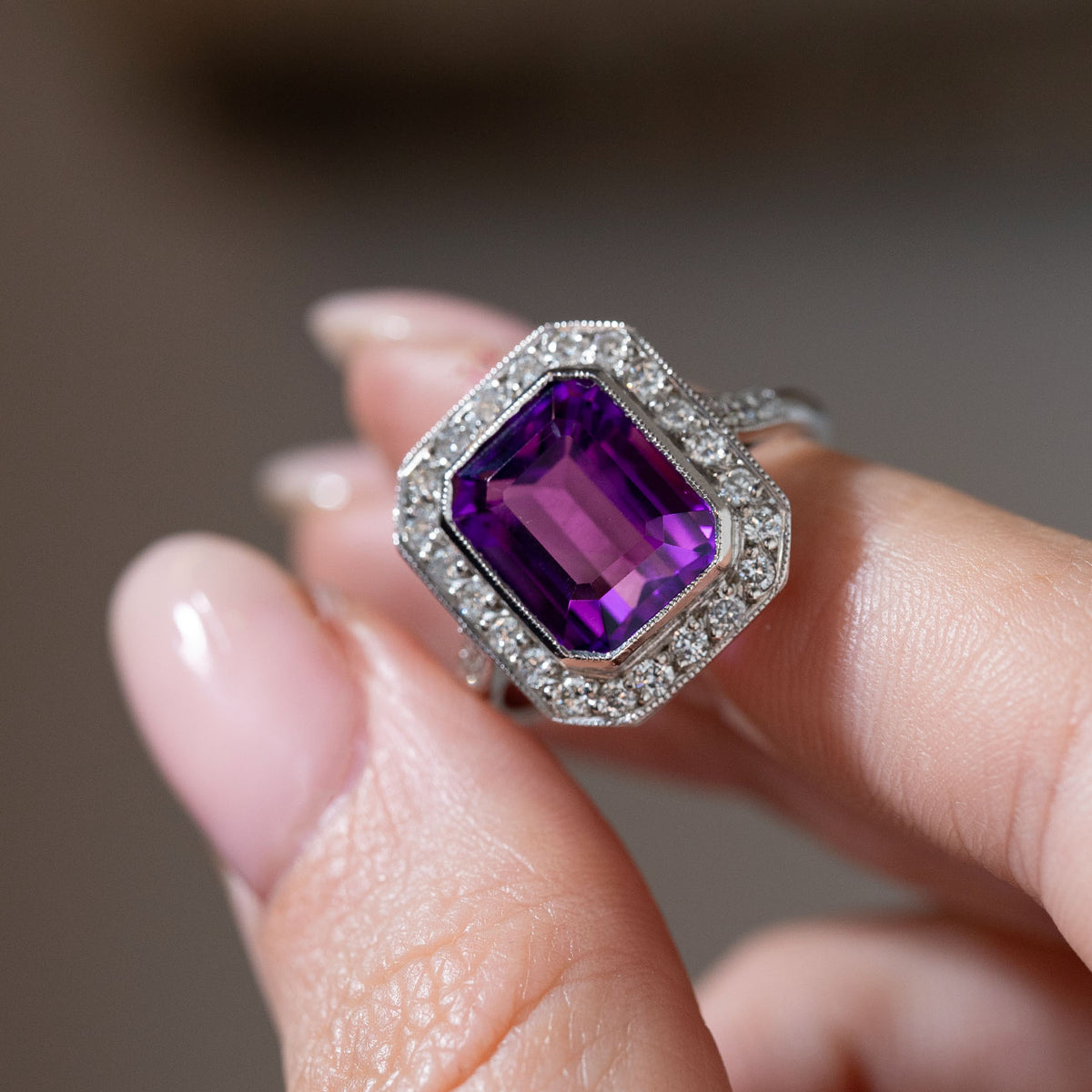 Detailed close-up of an antique Art Deco amethyst and diamond cluster ring held between fingers, highlighting the emerald-cut purple amethyst, octagonal diamond surround and intricate milgrain edges.