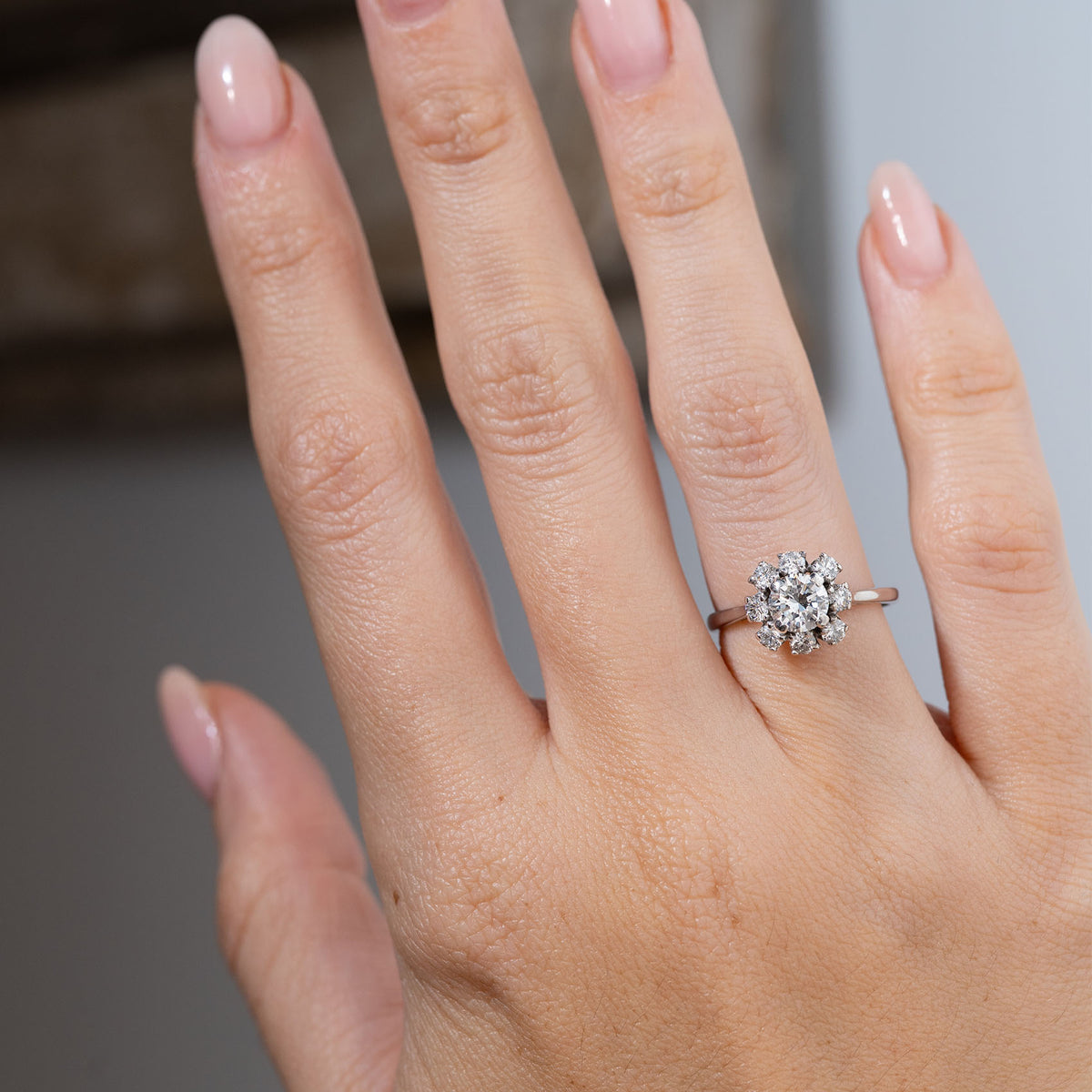 Vintage 1950s diamond cluster engagement ring worn on a hand, displaying its sparkling central diamond surrounded by eight smaller diamonds in a flower-shaped white gold setting.