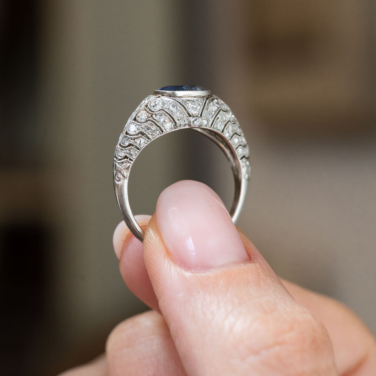 Reverse profile of an antique sapphire bombé ring held between the fingers, showing its curved platinum gallery and diamond-set shoulders.