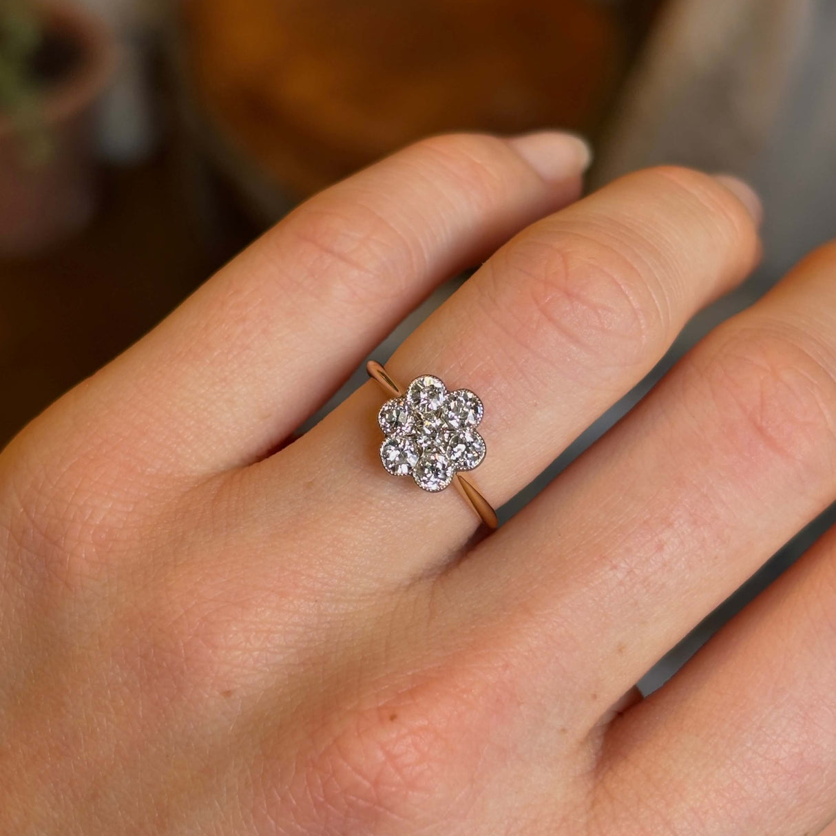 Close-up of a hand wearing an Edwardian daisy diamond cluster ring with an 18ct yellow gold band with a blurred background