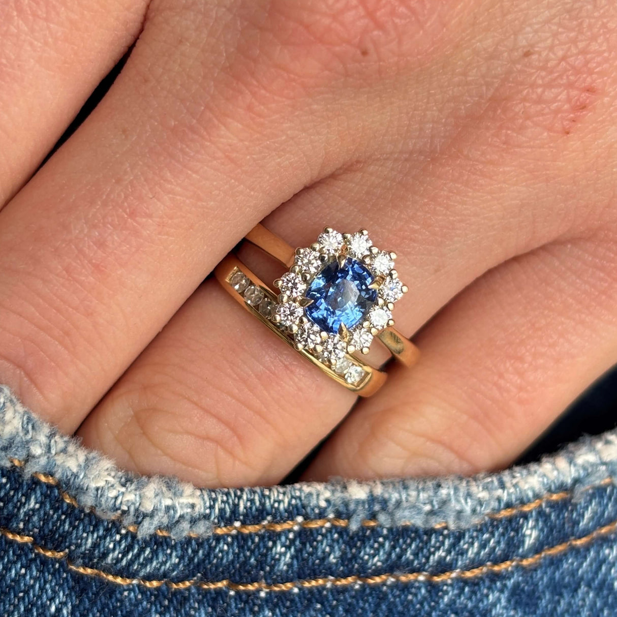 Blue sapphire and diamond cluster engagement ring stacked with a wedding band on hand in pocket of jeans.