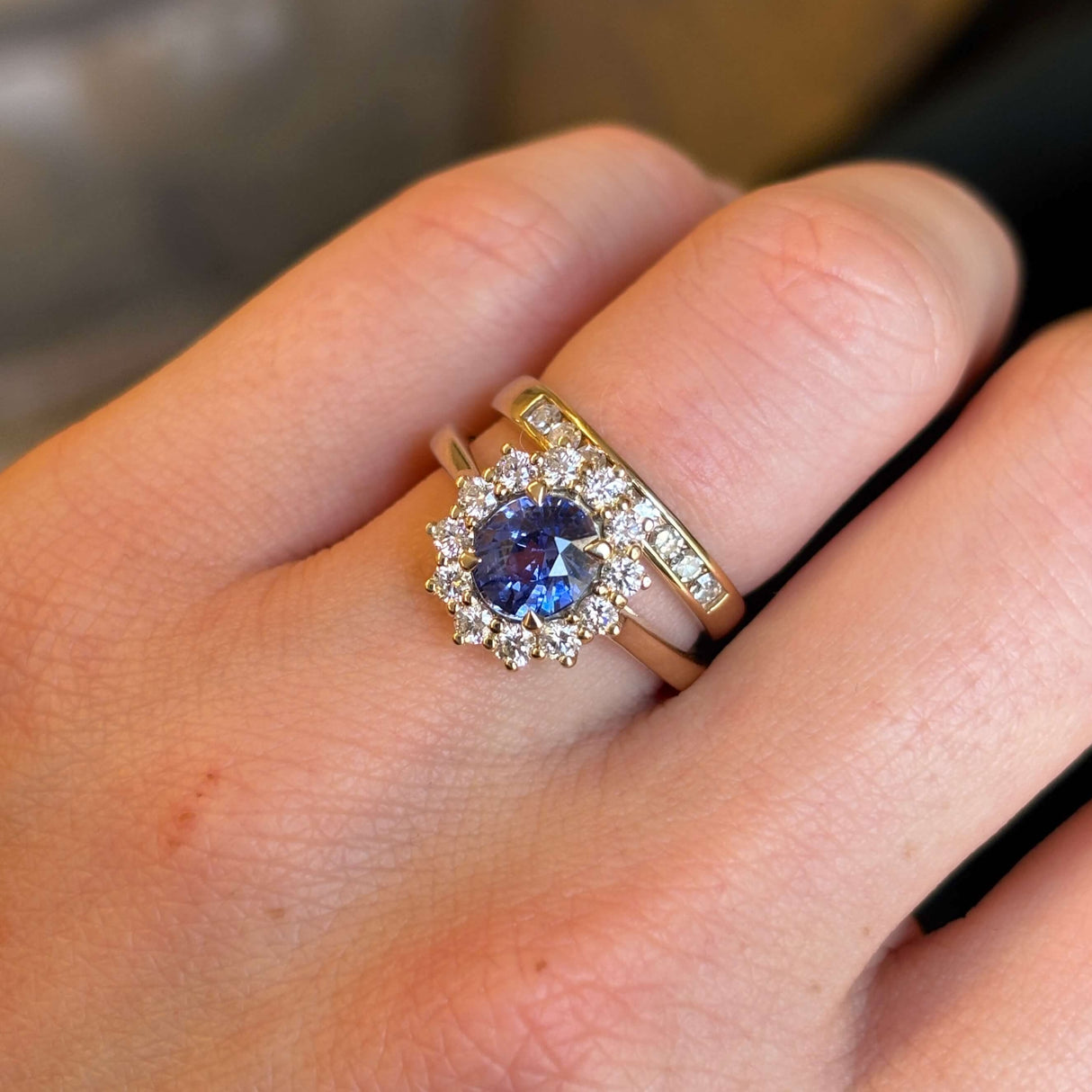 Blue sapphire and diamond cluster engagement ring worn on hand next to wedding band showing scale and proportion.