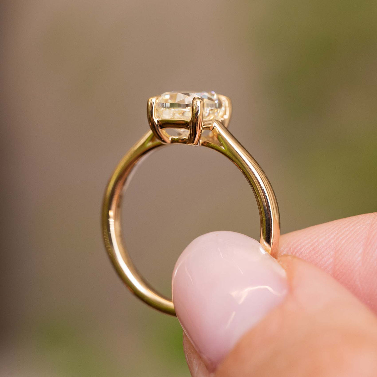 an image of an Antique-inspired, cushion-cut diamond solitaire engagement ring in yellow gold held up to show the profile of the ring against a blurred background