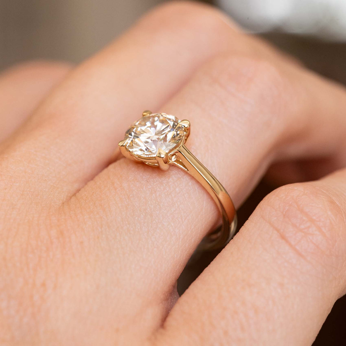 A close-up of a hand wearing a yellow gold solitaire engagement ring with a light brown diamond.