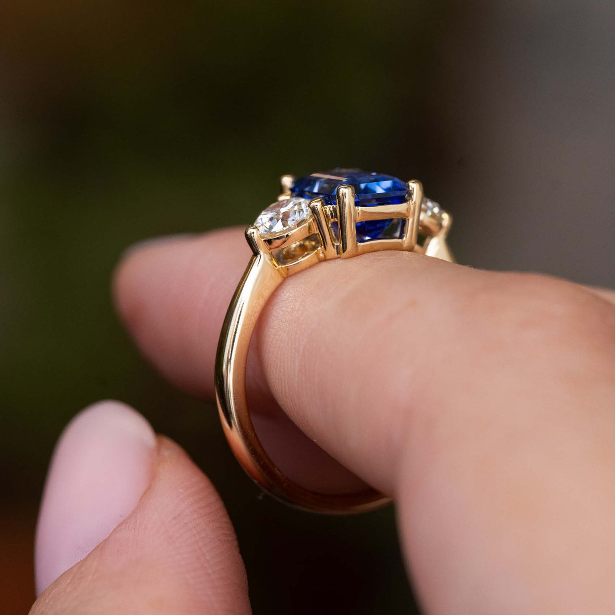 A close up side profile of an Antique-inspired, blue sapphire and diamond three-stone engagement ring, worn on a finger to show the low profile setting and yellow gold band.