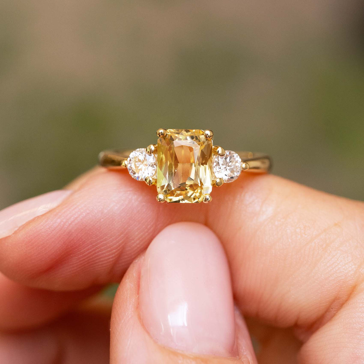 A close-up of a gold Antique-inspired, Yellow sapphire and diamond three-stone engagement ring balanced on the finger against a blurred natural background.