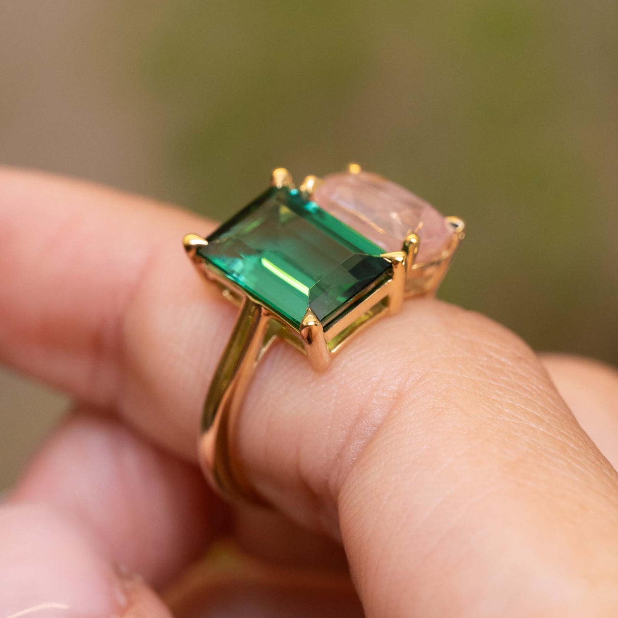 A yellow gold ring with a large emerald-cut green tourmaline and a pink sapphire worn on a person's finger against a blurred green background