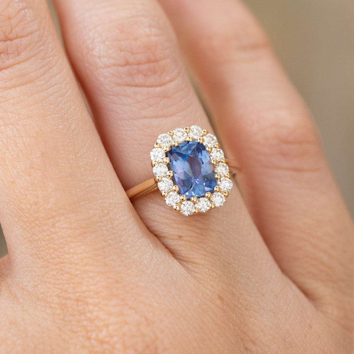 Antique-inspired sapphire engagement ring worn on the hand, showcasing its cushion-cut blue gemstone and sparkling round diamond halo.