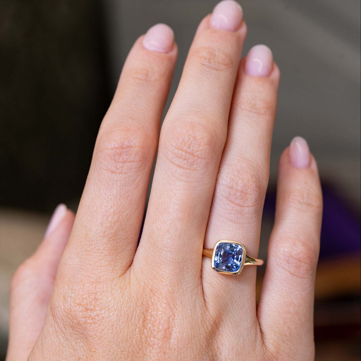 An image of an antique-inspired blue cushion cut sapphire bezel set in a yellow gold solitaire ring worn on the finger and shown at a distance against a blurred background.