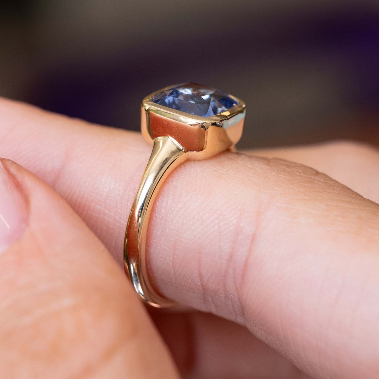 An image of an antique style blue sapphire single stone bezel-set yellow gold ring held on a finger and displayed from the side to show the setting style against a blurry background.