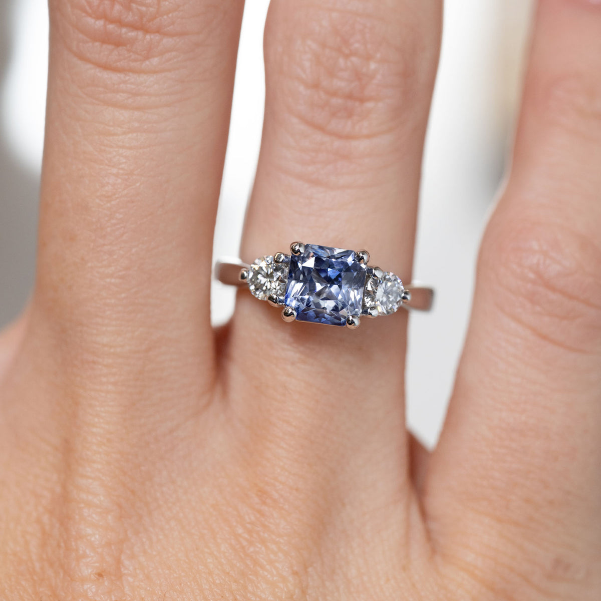 Close-up of the square cushion-cut blue sapphire flanked by two brilliant-cut white diamonds, worn on a finger.