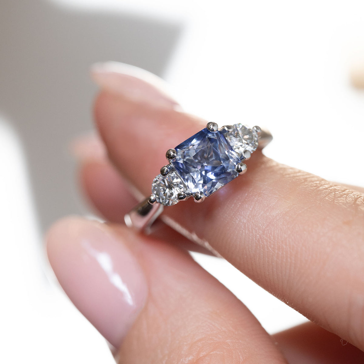 Hand-held close-up of the cushion-cut blue sapphire and round diamond shoulders in a polished platinum claw setting.