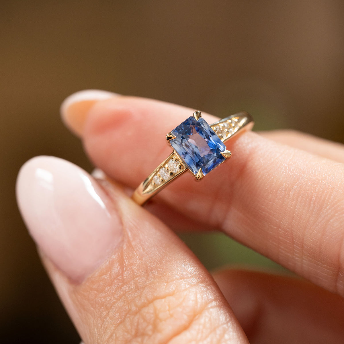 A close-up of a hand wearing a yellow gold Vintage-inspired, Blue sapphire and diamond flanked solitaire engagement ring set against a soft, blurred brown background.