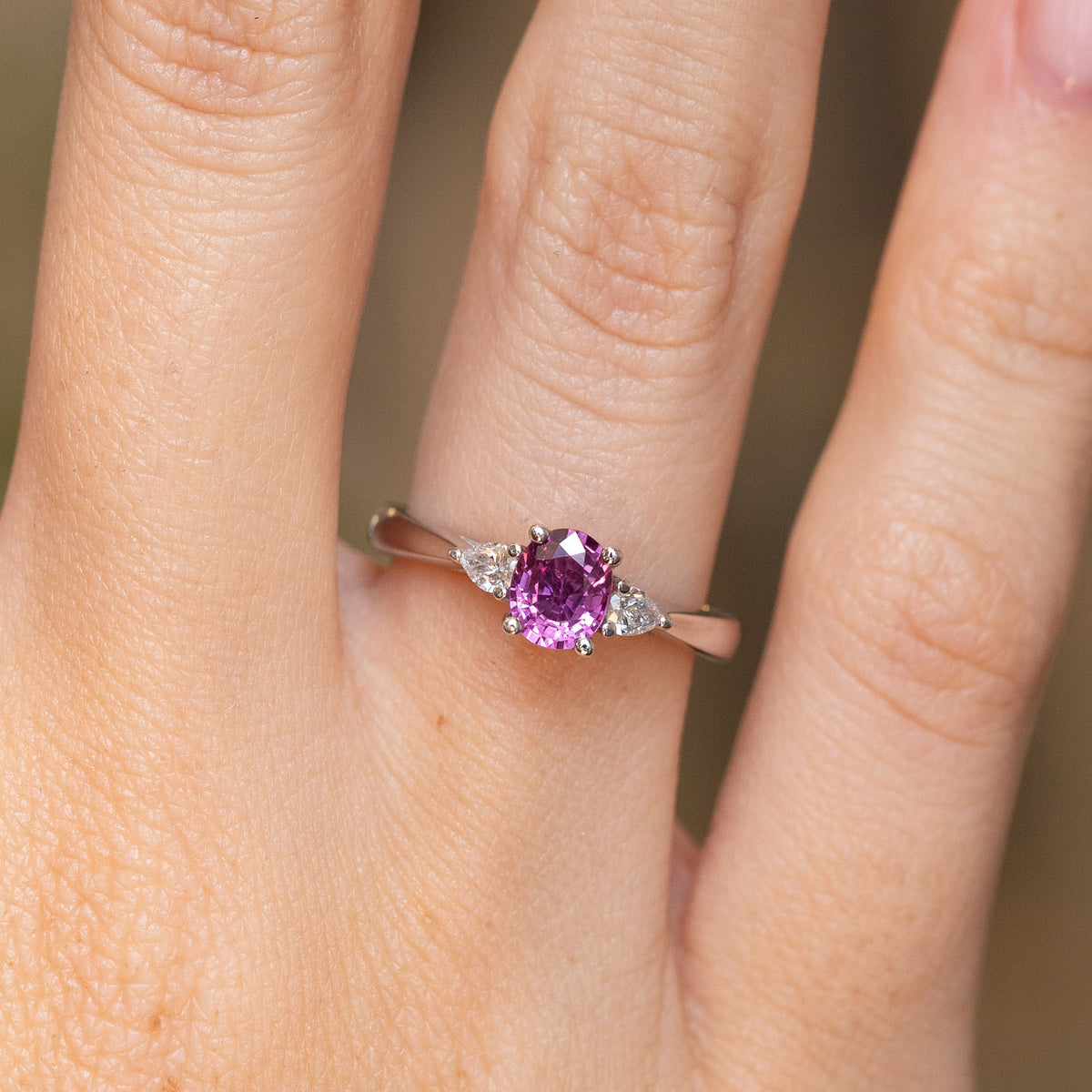 Close-up of an antique-style platinum engagement ring with a deep purple-pink sapphire between two sparkling pear-cut diamonds.