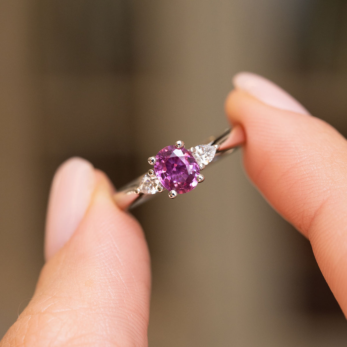 Antique-inspired three-stone engagement ring held between the fingers, with a vivid sapphire flanked by matching pear-shaped diamonds.