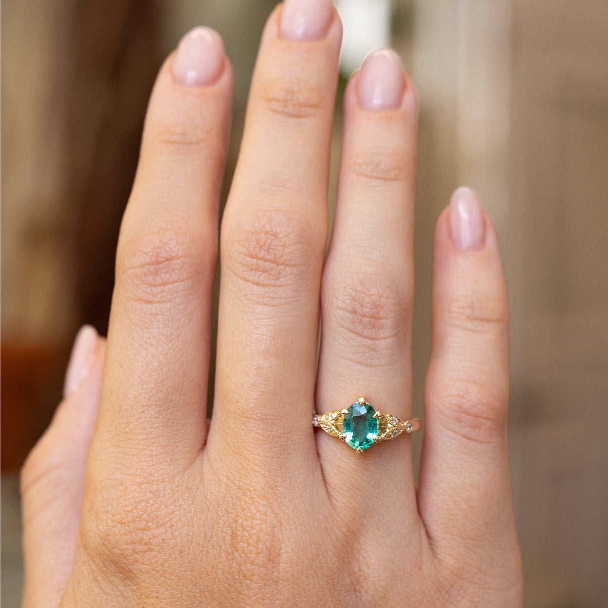 A close-up of a hand wearing a gold ring with a central oval emerald gemstone and small diamonds stones, set against a softly blurred warm brown background.