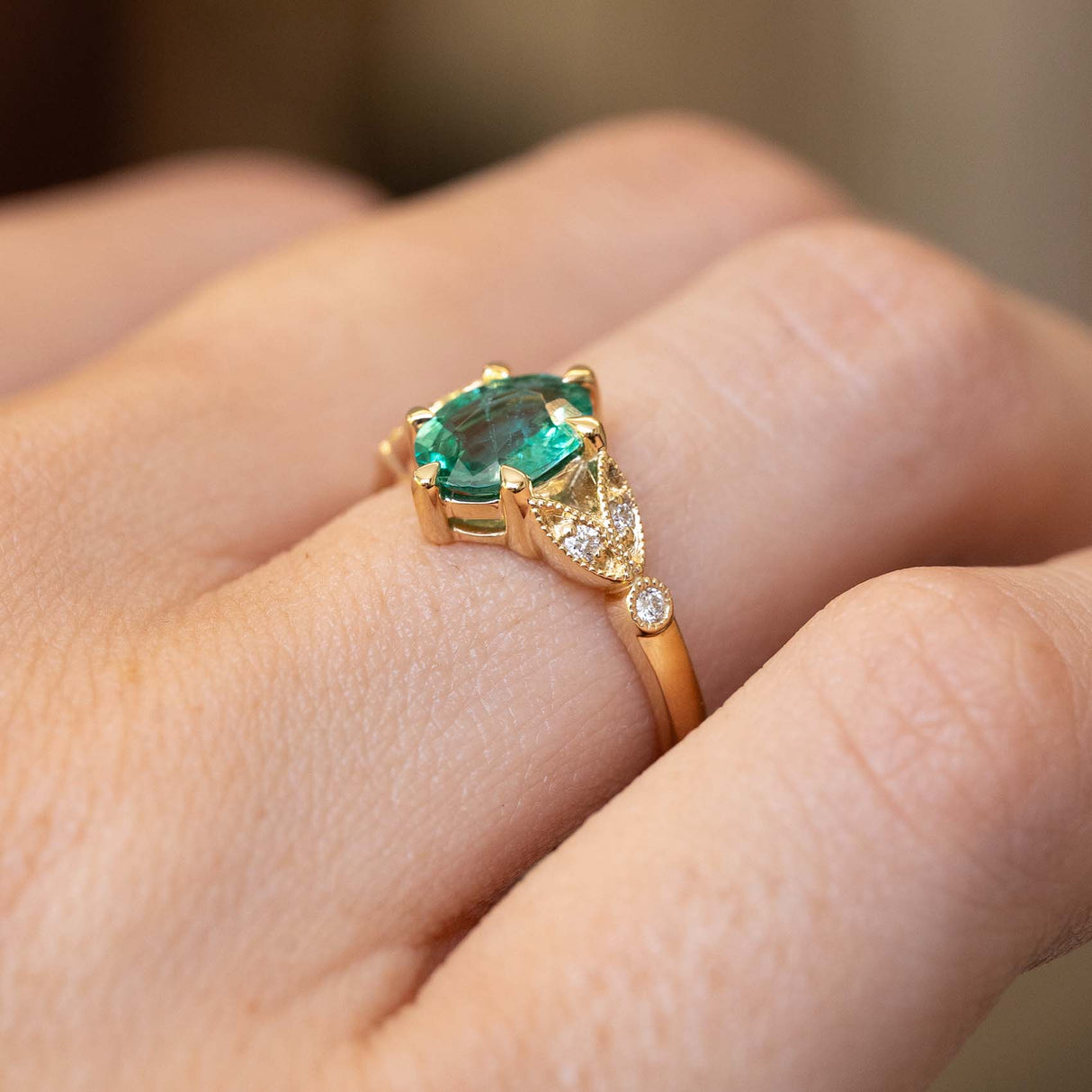 A close-up of a gold ring with a Vintage-inspired, Emerald and diamond engagement ring, worn on a person's finger.