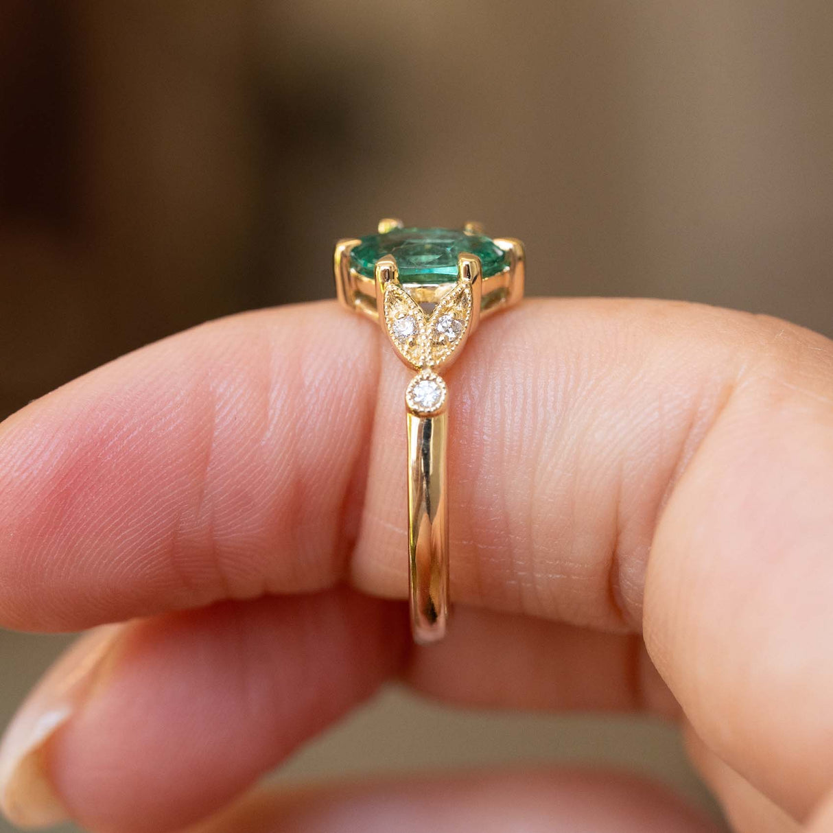A close-up of a gold ring with a green emerald trilogy engagement ring, worn on a person's finger against a blurred background.