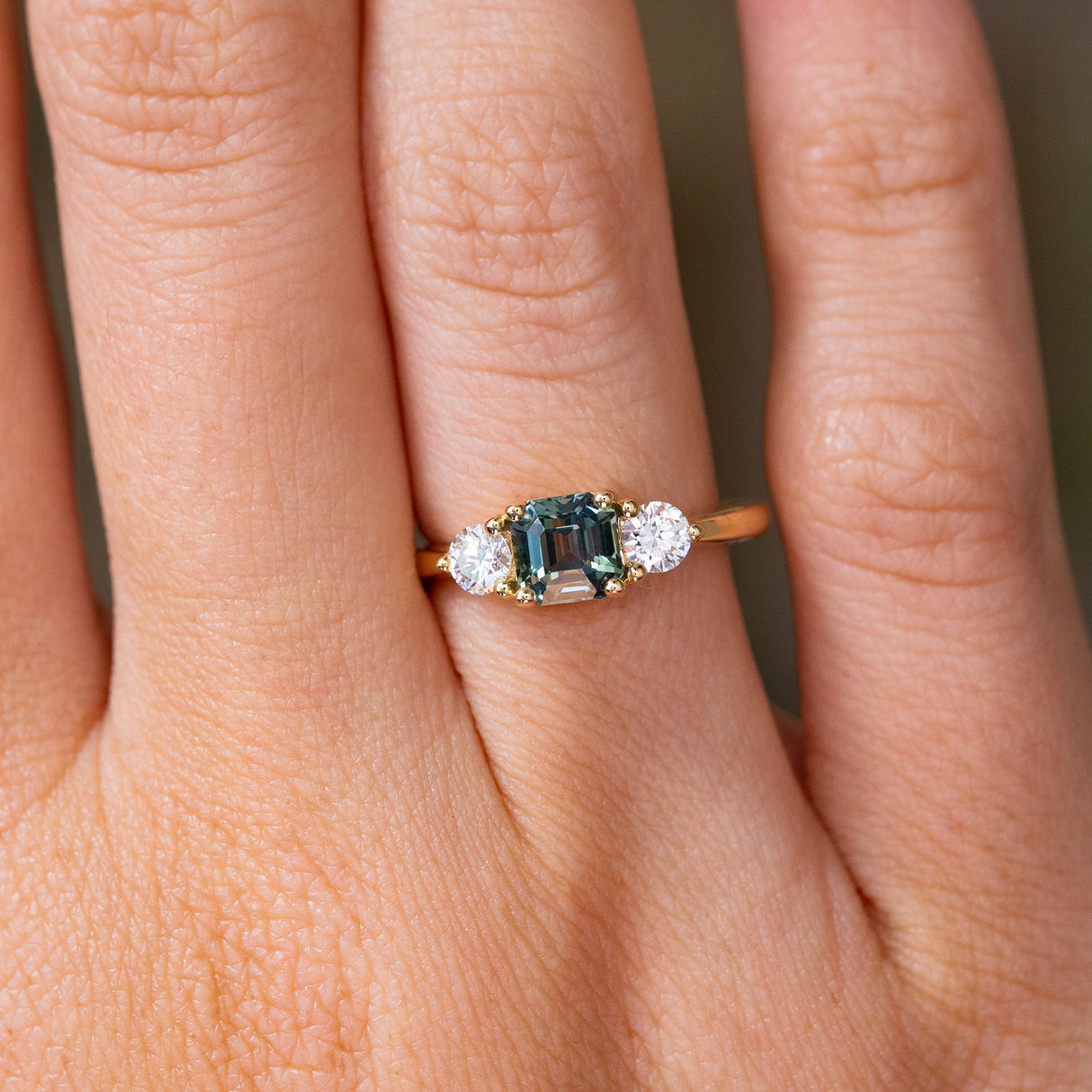 A close-up of a hand wearing a yellow gold Antique-inspired, Teal sapphire and diamond three-stone engagement ring