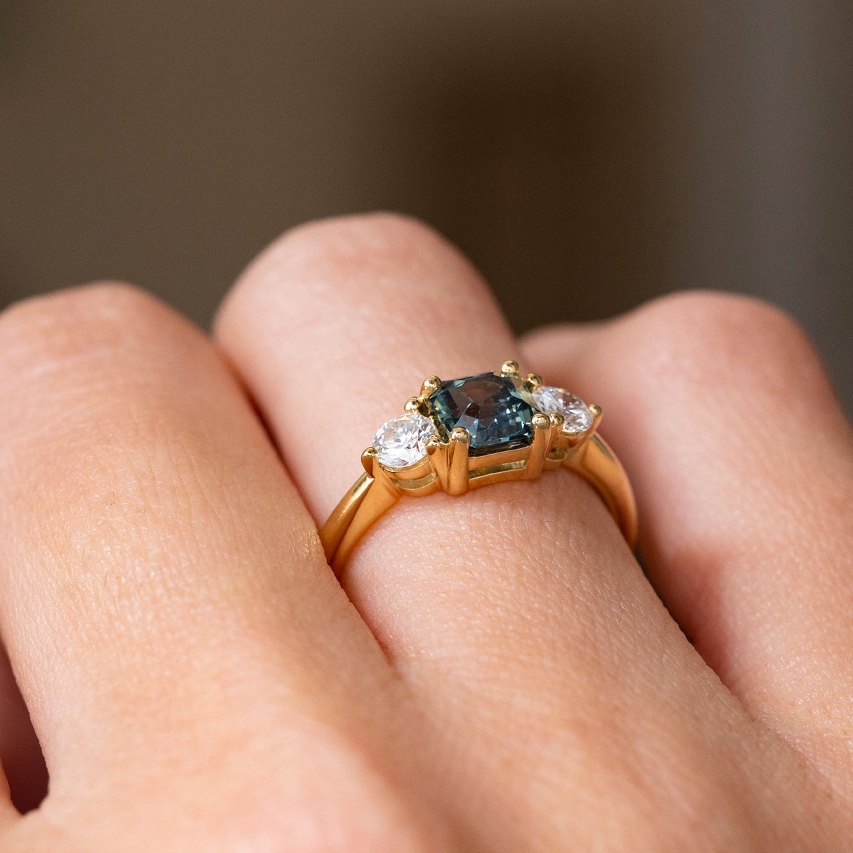 An Antique-inspired, Teal sapphire and diamond trilogy yellow gold engagement ring worn on the hand and shown from the side to display the gallery