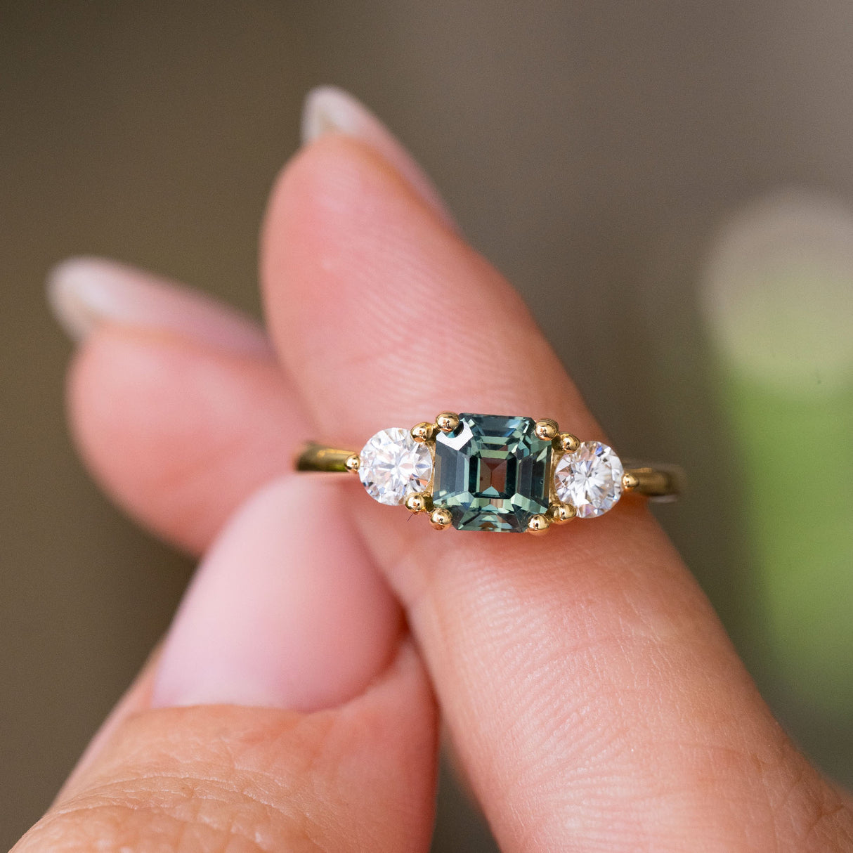 A close-up of a yellow gold Antique-inspired, Teal sapphire and diamond three-stone engagement ring balanced on the fingers and against a blurred background