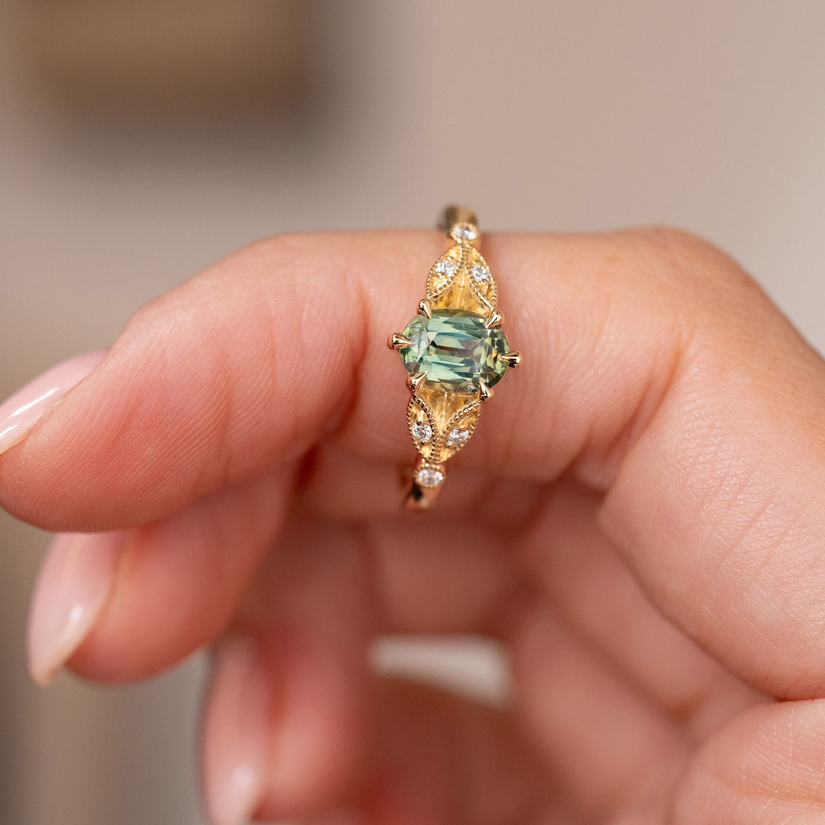 Teal sapphire and diamond ring held between the fingers, offering a clear view of its symmetrical vintage-inspired design.