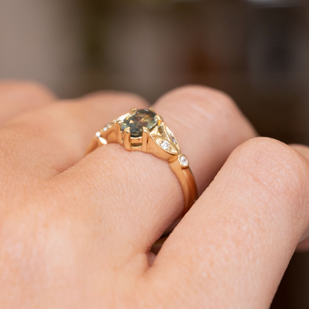 A close-up of a Vintage-inspired, Green sapphire and diamond engagement ring worn on a person's finger.