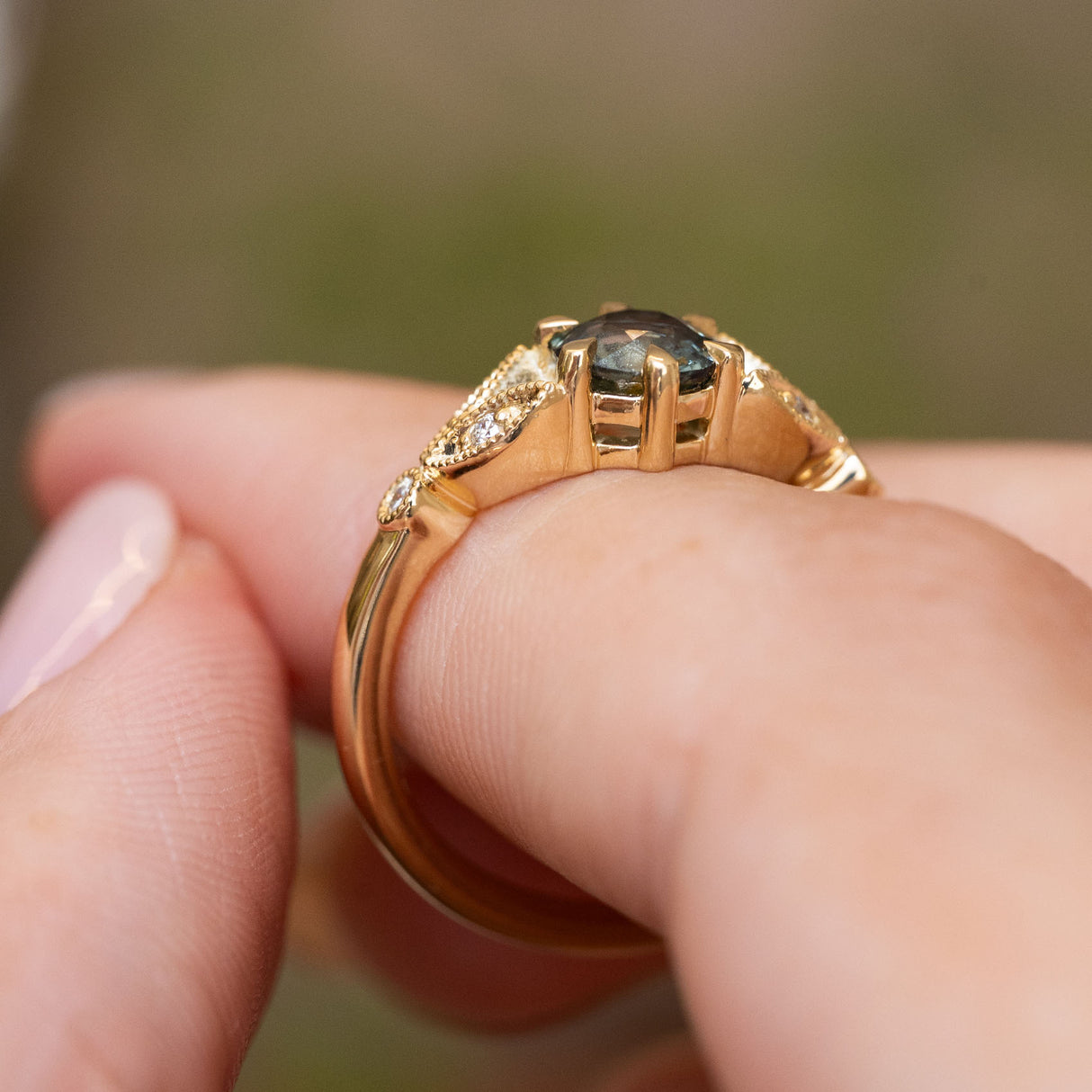 A close-up of the profile of a Vintage-inspired, Green sapphire and diamond engagement ring with a yellow gold band and claws.