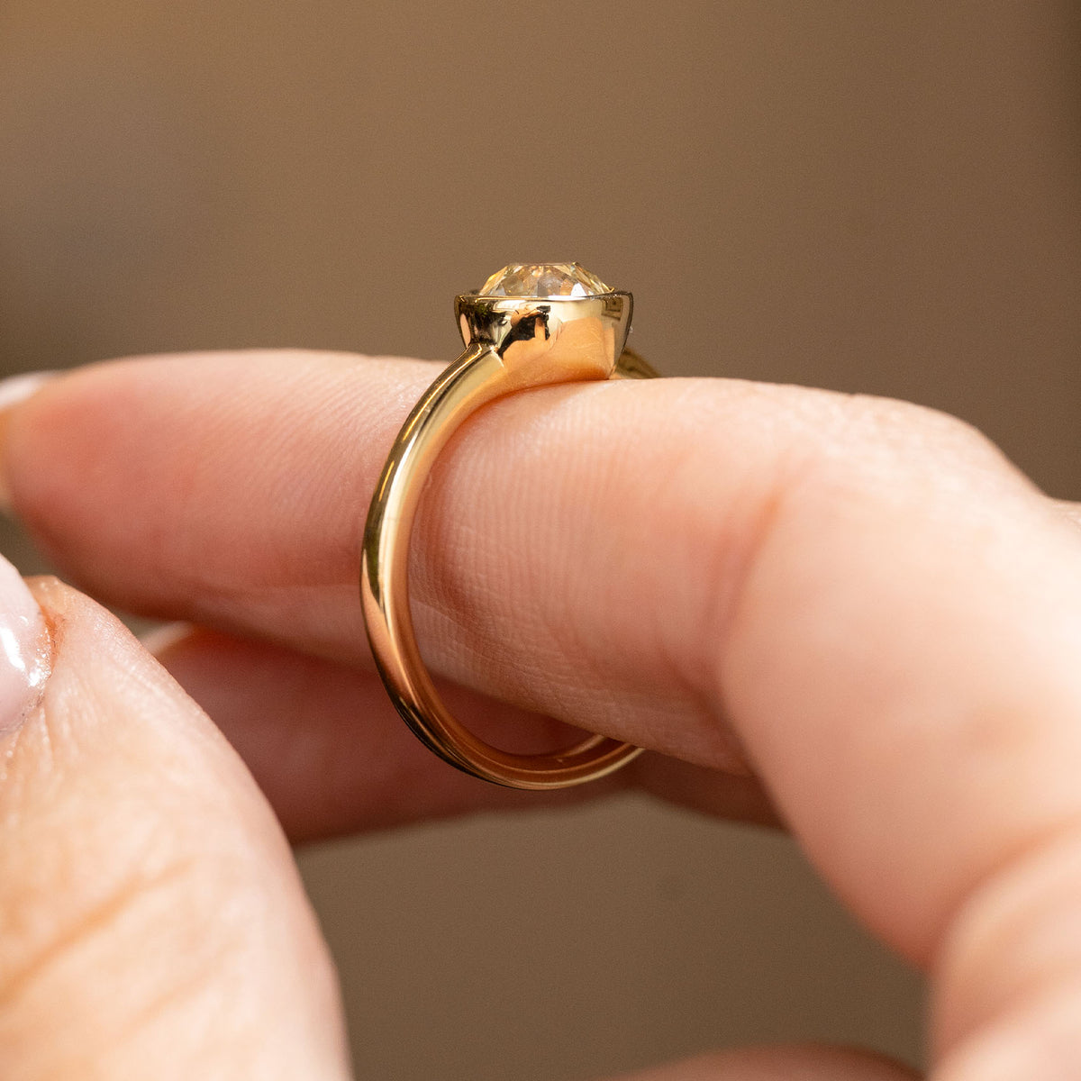 A close-up image of a Solitaire engagement ring in yellow gold with an old cut diamond basil set balanced on A close-up image of a solitaire engagement ring in yellow gold with an old cut diamond bezel set balanced on the finger and shown from a low side angle to showcase the shank and profile.