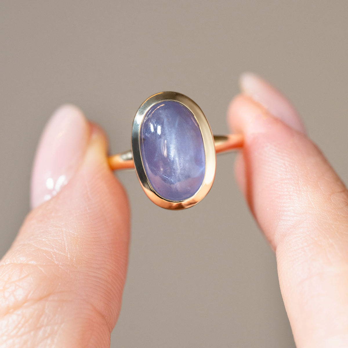 Detailed photograph of the oval cabochon sapphire held between fingers, highlighting its lavender-blue hues and natural cat's eye sheen.