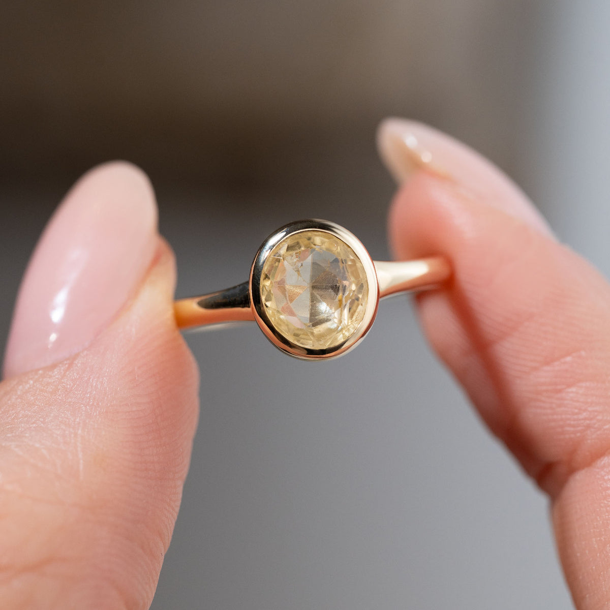 Close-up of an antique-inspired pale yellow sapphire solitaire engagement ring held between fingers, highlighting the soft yellow tones and delicate facets of the oval sapphire in its gold bezel setting.