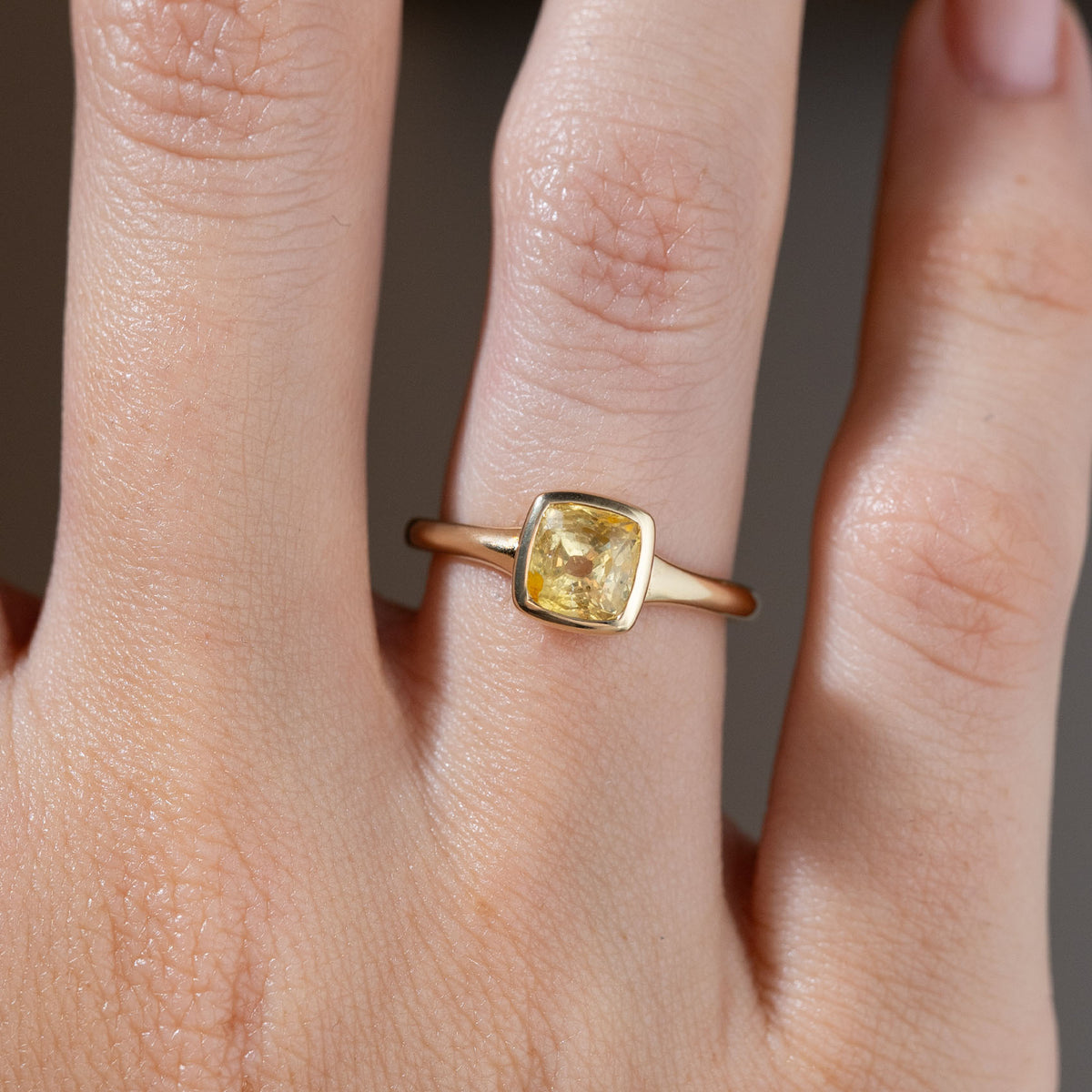 Close-up of the square cushion-cut yellow sapphire ring on a finger, showing its golden reflections and smooth bezel setting.