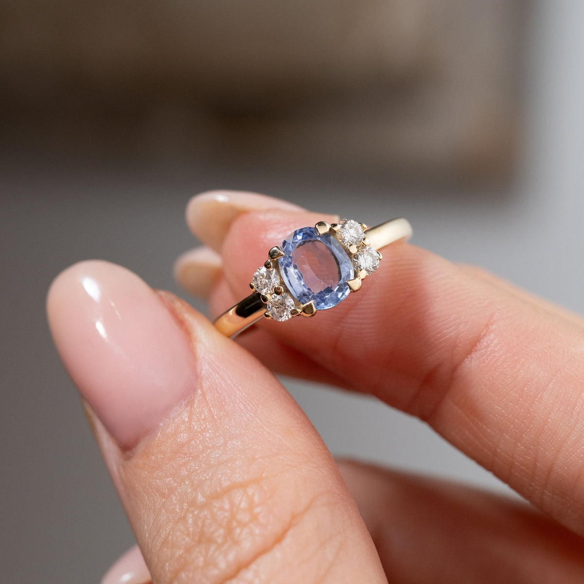 Close-up of the blue sapphire and diamond cluster engagement ring held between fingers, highlighting the sapphire's delicate blue reflections, surrounding diamonds and yellow gold claw setting.