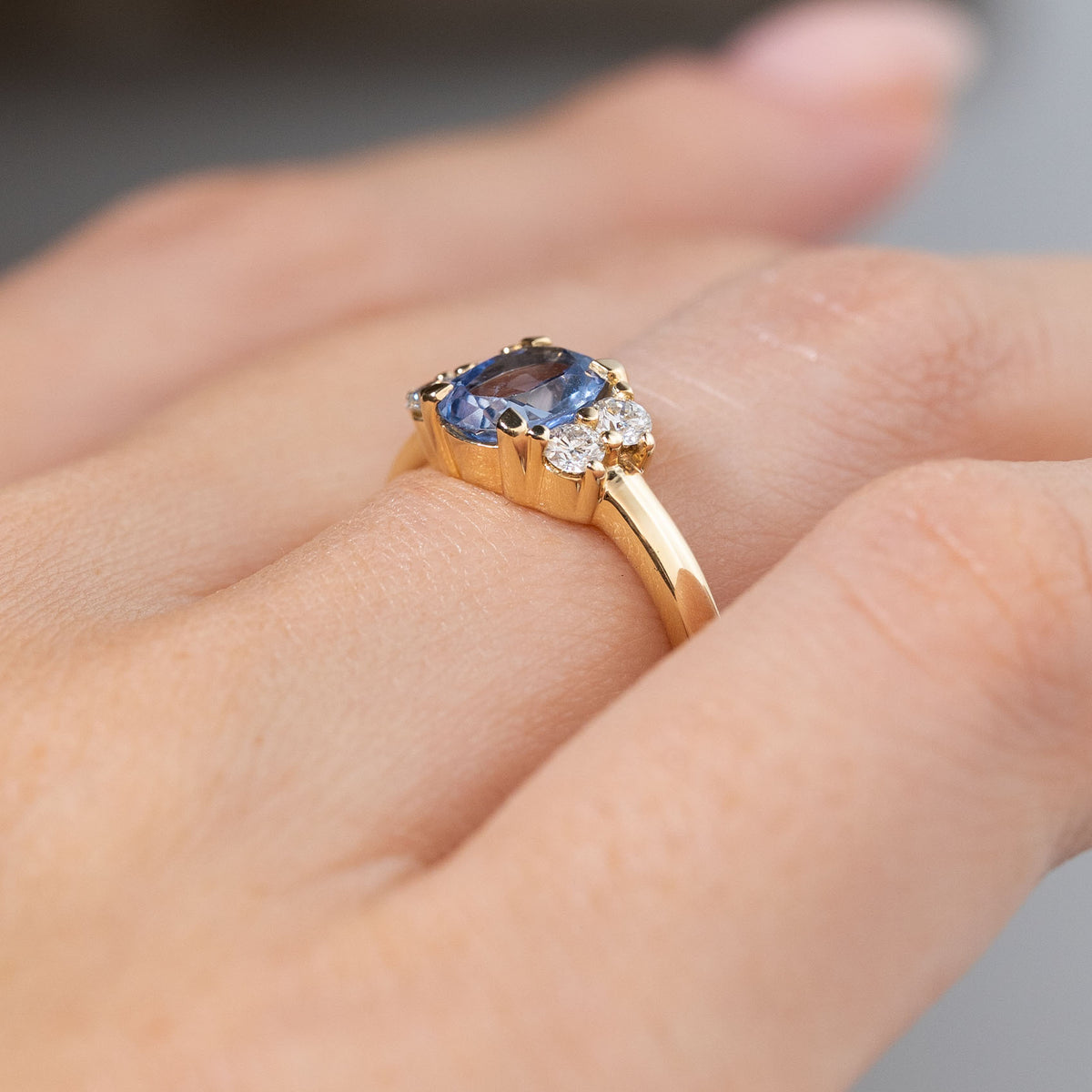 Angled side view of the vintage-inspired blue sapphire and diamond cluster engagement ring worn on a hand, showcasing its raised claw setting, sparkling diamonds and slender yellow gold band.