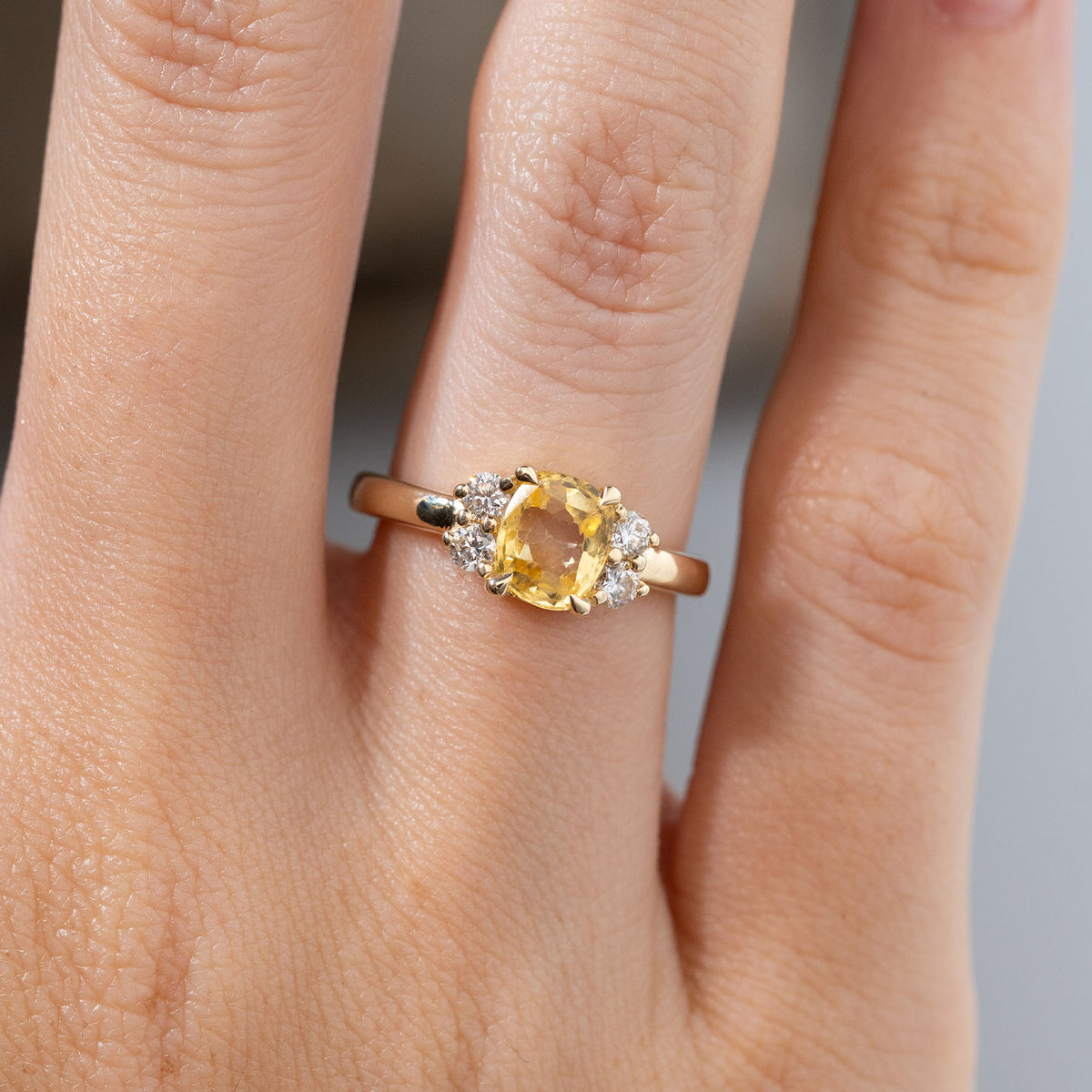 Close-up of the cushion-cut yellow sapphire engagement ring worn on a finger, showcasing its vibrant golden colour, four surrounding diamonds and polished yellow gold band.