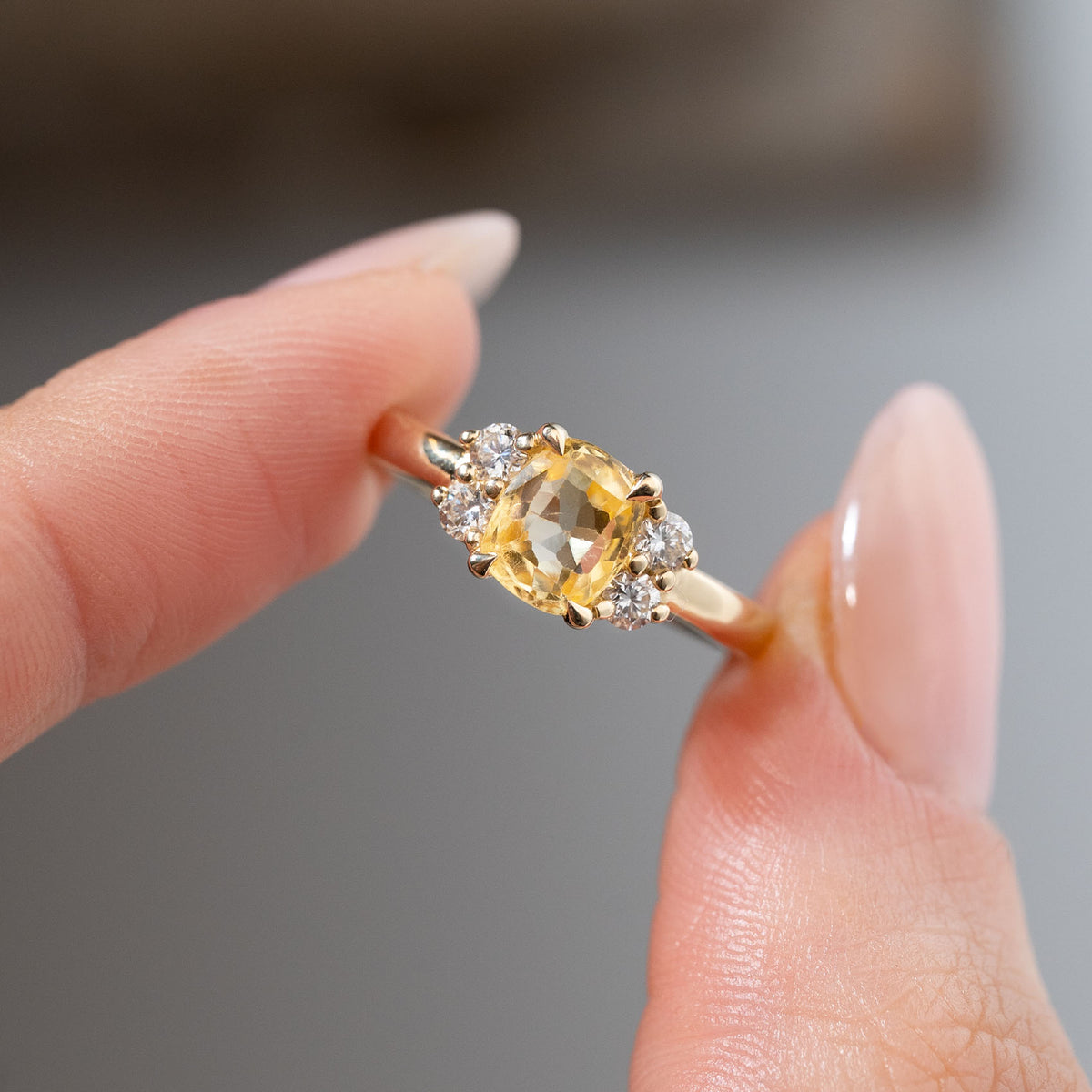 Detailed close-up of the vintage-inspired yellow sapphire and diamond engagement ring held between fingers, highlighting the cushion-cut gemstone, symmetrical diamond arrangement and polished gold claws.