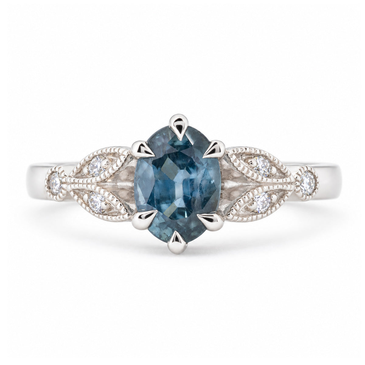 Front view of a vintage-inspired oval-cut sapphire engagement ring in platinum, featuring a muted blue gemstone and decorative diamond-set trefoil shoulders inspired by laurel leaves.